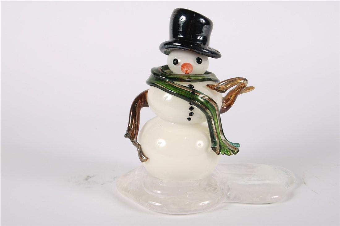 Art Glass Snowman: Glass, 2013. Height: 9 1/2", Length: 9", Depth: 5 1/2". White body with clear glass base speckled with white. Green flowing scarf, black top hat. Signature to upper side of base. Signature looks like 