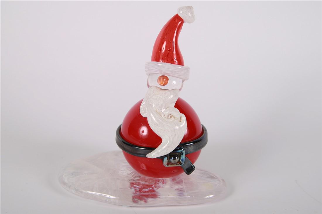 Art Glass Snowman in Santa Suit: Glass. Height: 8 1/4", Length: 7", Depth: 4 3/4". Whimsical depiction of snowman in a red santa suit and hat, with a clear base swirled with white. Signature to edge of base.