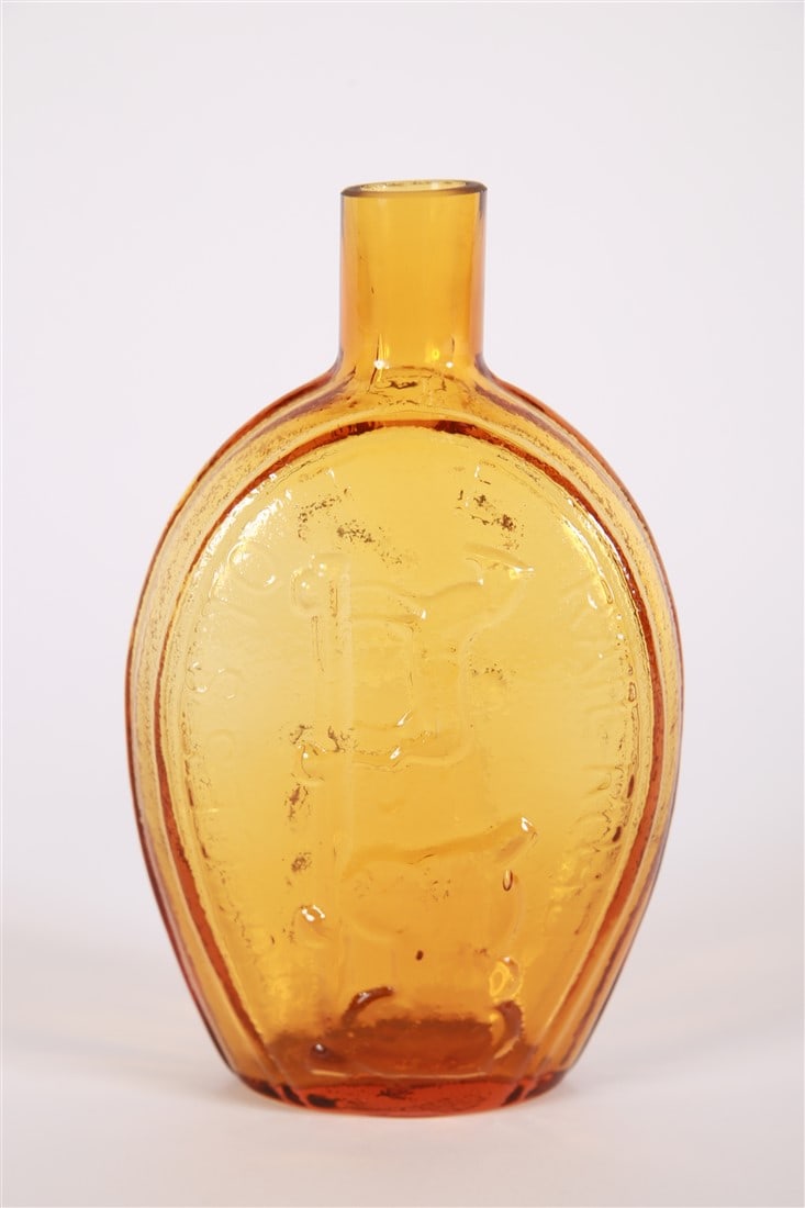 "Success to the Railroad" Glass Flask: Glass. Height: 7", Length: 4 1/4", Depth: 2 1/4". Amber colored glass flask. Both sides with an image of a horse and cart on a railway track and reads, "Success to the Railroad".