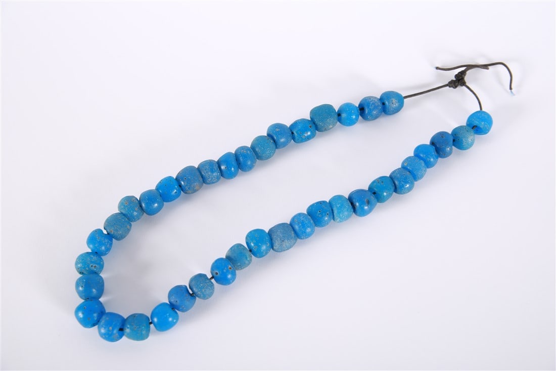 Blue Glass Trade Bead Necklace: Glass, leather cord. Length of necklace: 26". Strand of beads comprised of spherical blue glass on a black leather cord. Irregular shape to beads.