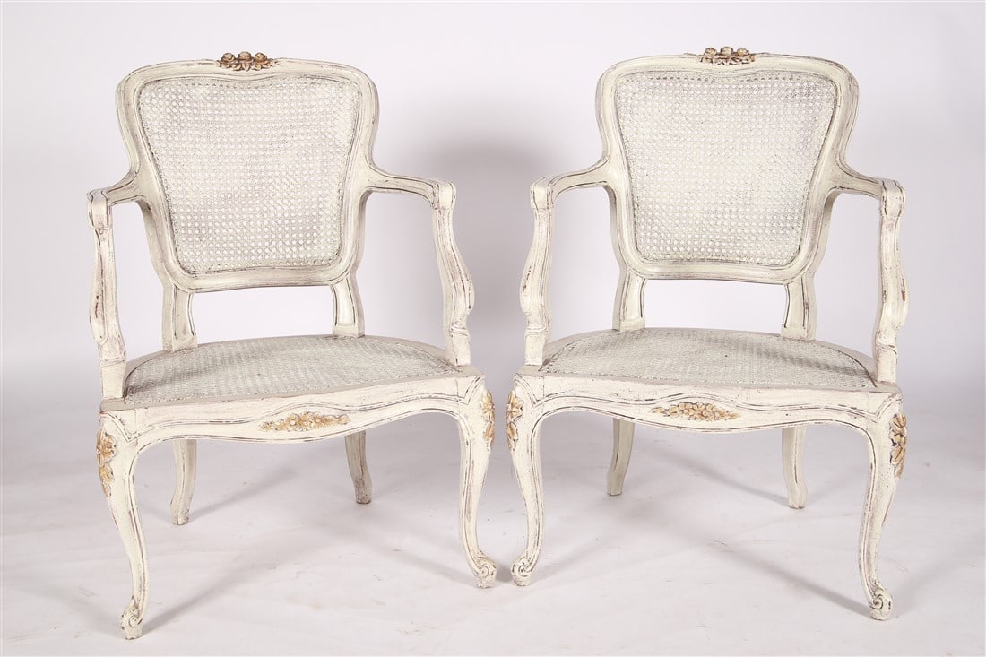 Pair of French Louis XV Style Caned Armchairs: Wood, cane. Height: 32", Length: 22", Depth: 19 1/2". Matching pair of Louis XV style armchairs with woven cane and a distressed white painted finish. Gilded floral details on front and legs.