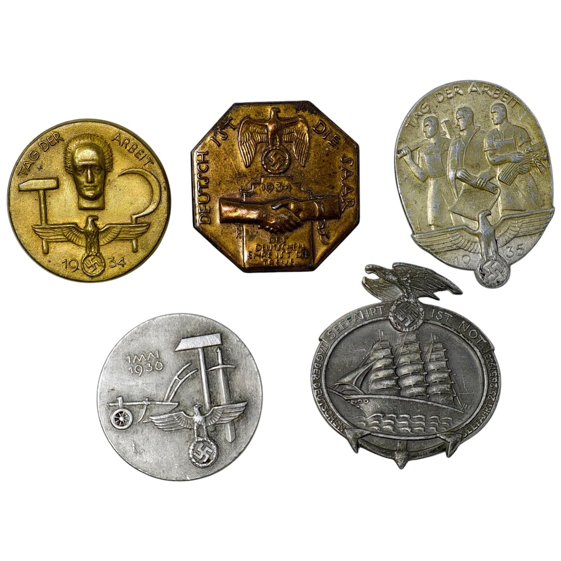 Third Reich Nazi Set of Tinnie Badges: A set of pin-back tinnie badges, including a set commemorating Tag der Arbeit (May Day), one marked "Deutsch Ist Die Saar" 1934 commemorating the Saar reunification, and one nautical themed "Seefahrt 