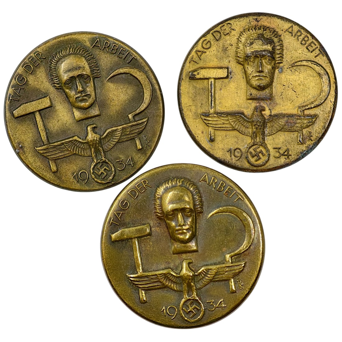 Third Reich 'Tag der Arbeit' 1934 Tinnie Badge Set: A set of pin badges in what appears to be a brass/bronze tone commemorating "Tag der Arbeit" (National Labor Day), dated 1934. Each depicts a stylized worker's head flanked by a hammer and sickle