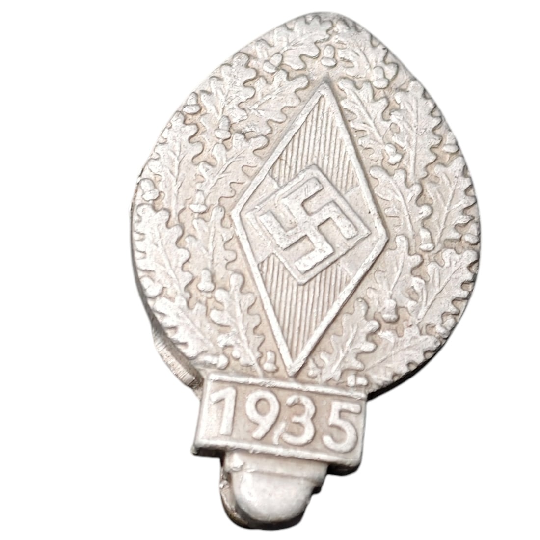 Hitler Youth 1935 Pin: A teardrop-shaped pin badge featuring a central diamond with swastika motif surrounded by an oak leaf wreath, with a dated tab reading '1935' at the base. Appears to be cast from a metal that appears 