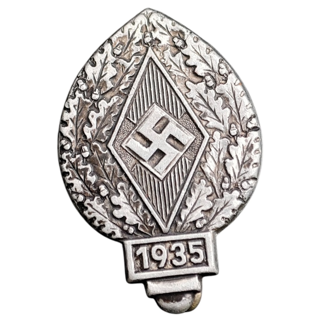 Hitler Youth Event Badge 1935: A leaf-shaped tinnie badge commemorating a 1935 Hitler Youth (Hitlerjugend) event, featuring a diamond-shaped HJ emblem framed by oak leaves and acorns, with '1935' at the base. The reverse is maker-m