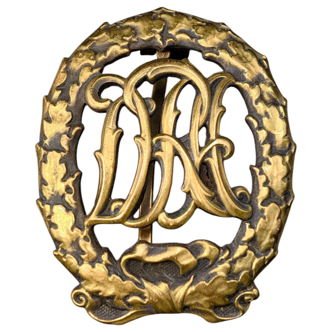 German DRL Badge in Bronze: A badge in what appears to be bronze-toned German Deutsches Reichsabzeichen für Leibesübungen (DRL) sports badge, featuring an oak leaf wreath surrounding the stylized 'DRL' monogram. The reverse is