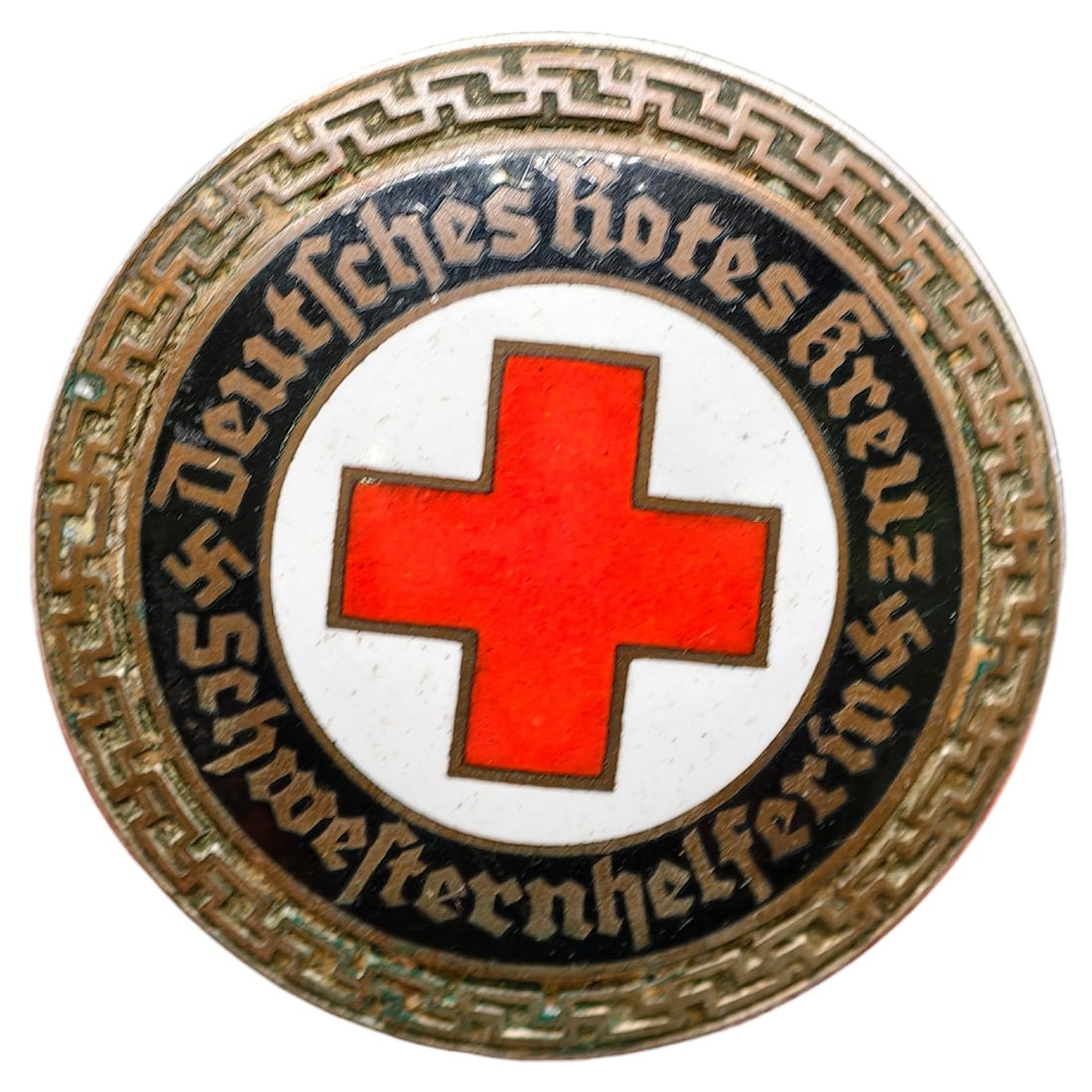 Third Reich Red Cross Badge: Round enameled badge inscribed "Deutsches Rotes Kreuz Schwesternhelferin" surrounding a central red enamel cross on white ground, bordered by a stylized meander/key pattern. Reverse stamped "GES. GESC