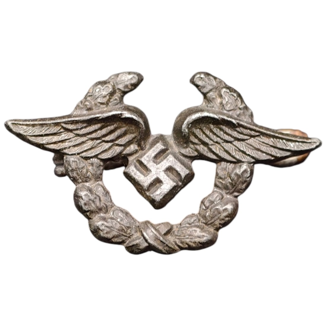 Third Reich Nazi Civil Employees Badge of the Luftwaffe: A small metal insignia depicting spread wings flanking a central swastika, encircled by an oak leaf wreath. No visible maker marks are present. The piece appears to be a cast white metal alloy with a 