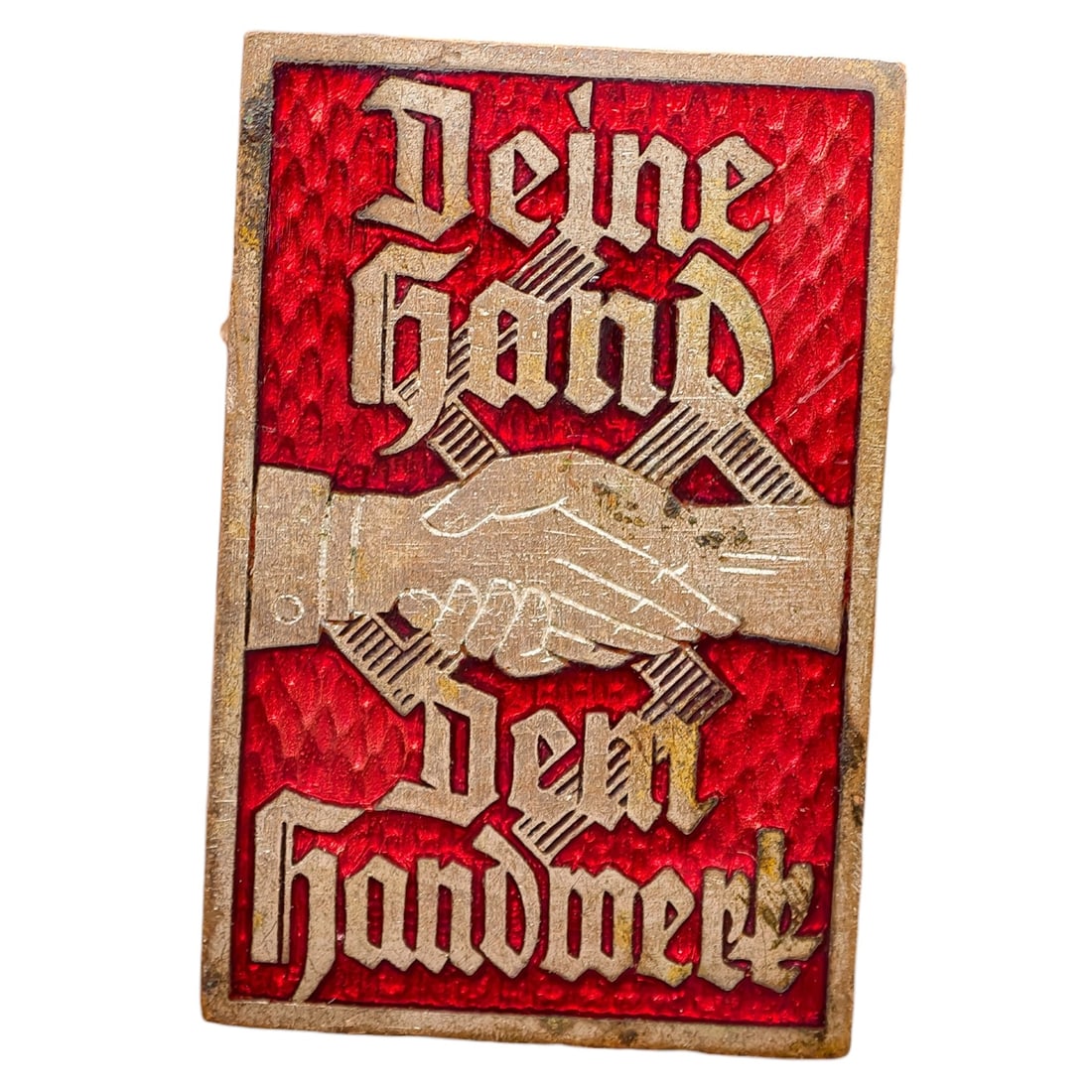 Third Reich Nazi Handwerk Badge: A stamped metal tinnie badge featuring the slogan 'Deine Hand Dem Handwerk' (Your Hand for the Trade/Craft) in Gothic script, with a central clasped-hands motif set against a textured red enamel-paint