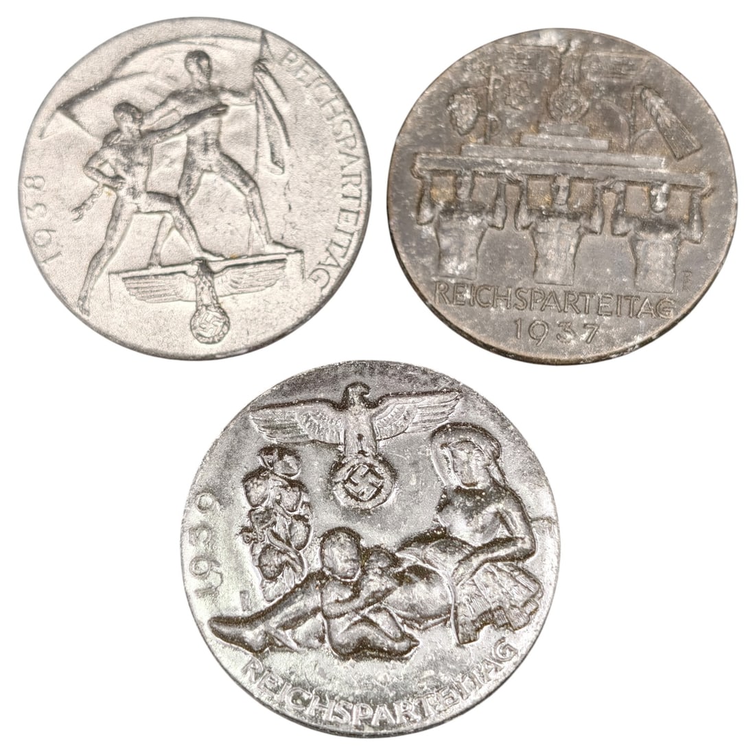 Third Reich Nazi Set of Tinnie Badges: A set of pin-back membership/day badges ("Tinnies") commemorating the Nuremberg Rally (Reichsparteitag) of 1937, 1938, and 1939. Each is die-struck in what appears to be white or gray metal with