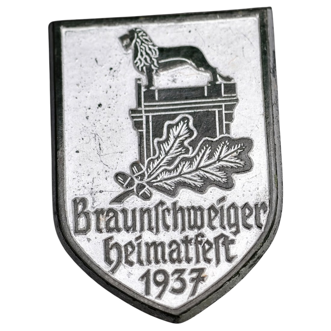 German 1937 Braunschweiger Heimatfest Tinnie Badge: A shield-shaped commemorative badge (tinnie) issued for the 1937 Braunschweiger Heimatfest, depicting the Brunswick Lion atop a plinth with an oak leaf and acorn spray below. Inscribed 'Braunschweiger