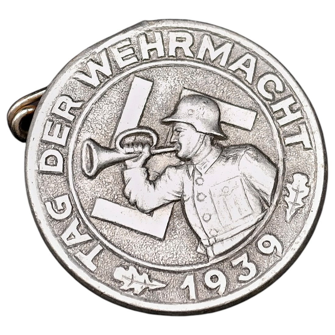 German WWII "Tag der Wehrmacht 1939" Commemorative Tinnie Badge: A pin-back commemorative badge issued for "Tag der Wehrmacht" (Day of the Armed Forces) 1939, depicting a helmeted soldier sounding a bugle flanked by stylized flags. Struck in a lightweight white met