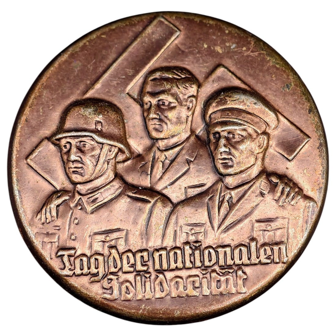 Third Reich Tag der Nationalen Soldacitat Tinnie Badge: A copper/bronze-toned commemorative tinnie depicting a set of figures - a Wehrmacht soldier in helmet, a civilian party member, and a worker in cap - shoulder to shoulder before crossed banner devices