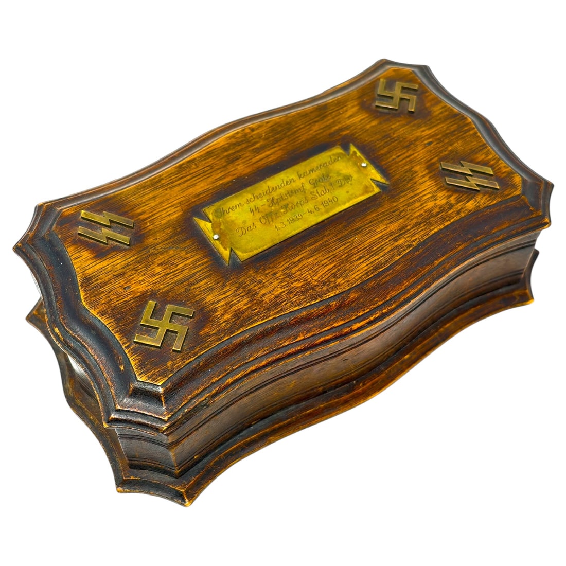 Third Reich SS Decorative Wood Box: A shaped wooden presentation box with a scalloped molded edge, the lid inlaid with a set of applied brass fittings: swastikas and SS sig-rune emblems. The center of the lid bears an engraved brass pla