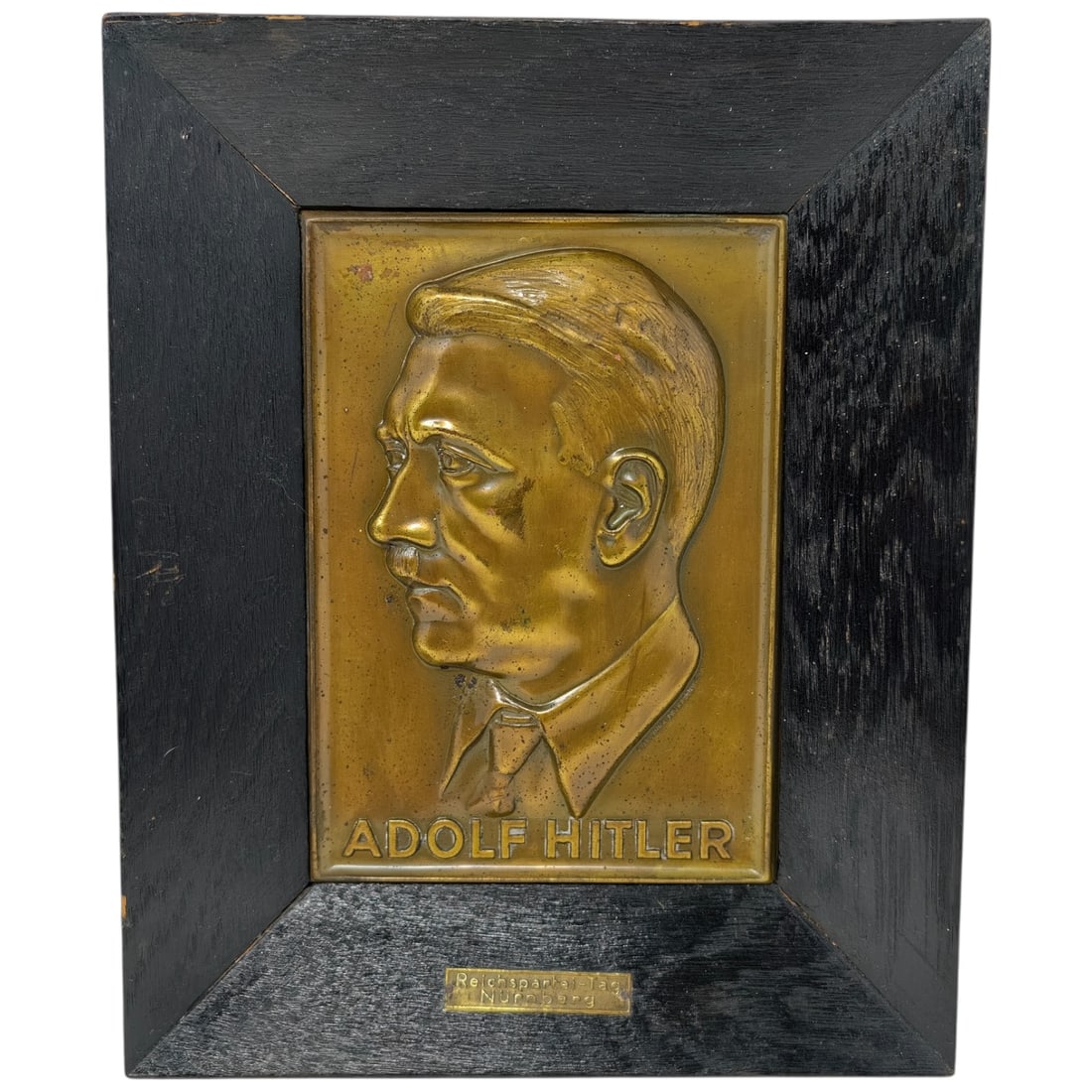 Adolf Hitler Reichsparteitag Nuremberg Plaque: A cast metal relief plaque depicting a left-facing profile portrait of Adolf Hitler above the raised lettering "ADOLF HITLER," mounted in a stepped wooden frame. A small metal tag affixed to the frame