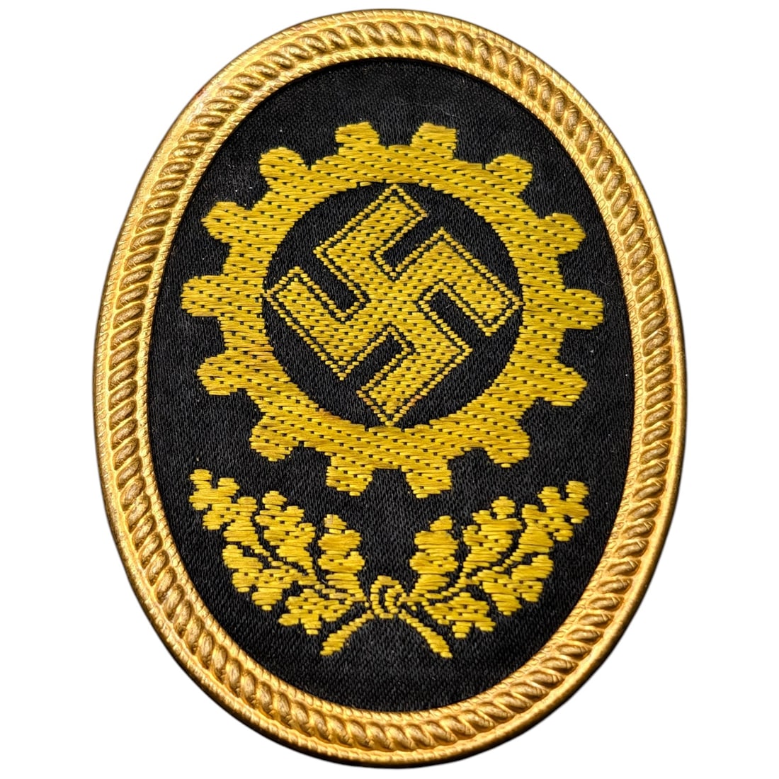 Third Reich Nazi DAF Insignia Badge: A bullion/wire-embroidered gear-and-cross insignia associated with the Deutsche Arbeitsfront (DAF), with oak leaf wreath below, mounted in an oval frame in what appears to be gilt metal. The reverse b