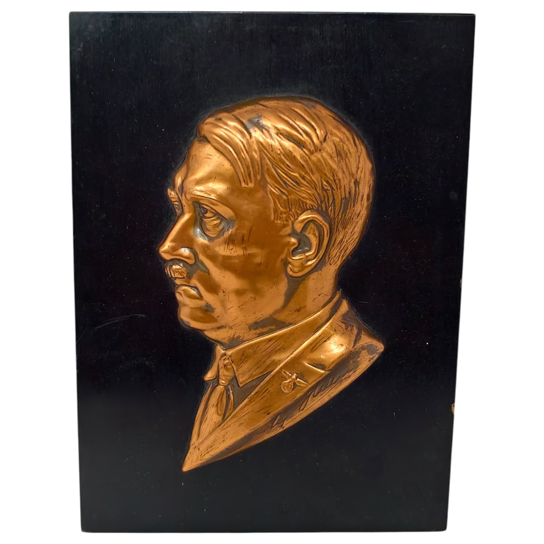 Adolf Hitler Wall Plaque: A relief-embossed copper portrait plaque depicting a profile bust, mounted on a black-painted wooden panel. A facsimile signature appears below the collar, and a small Party pin/eagle motif is