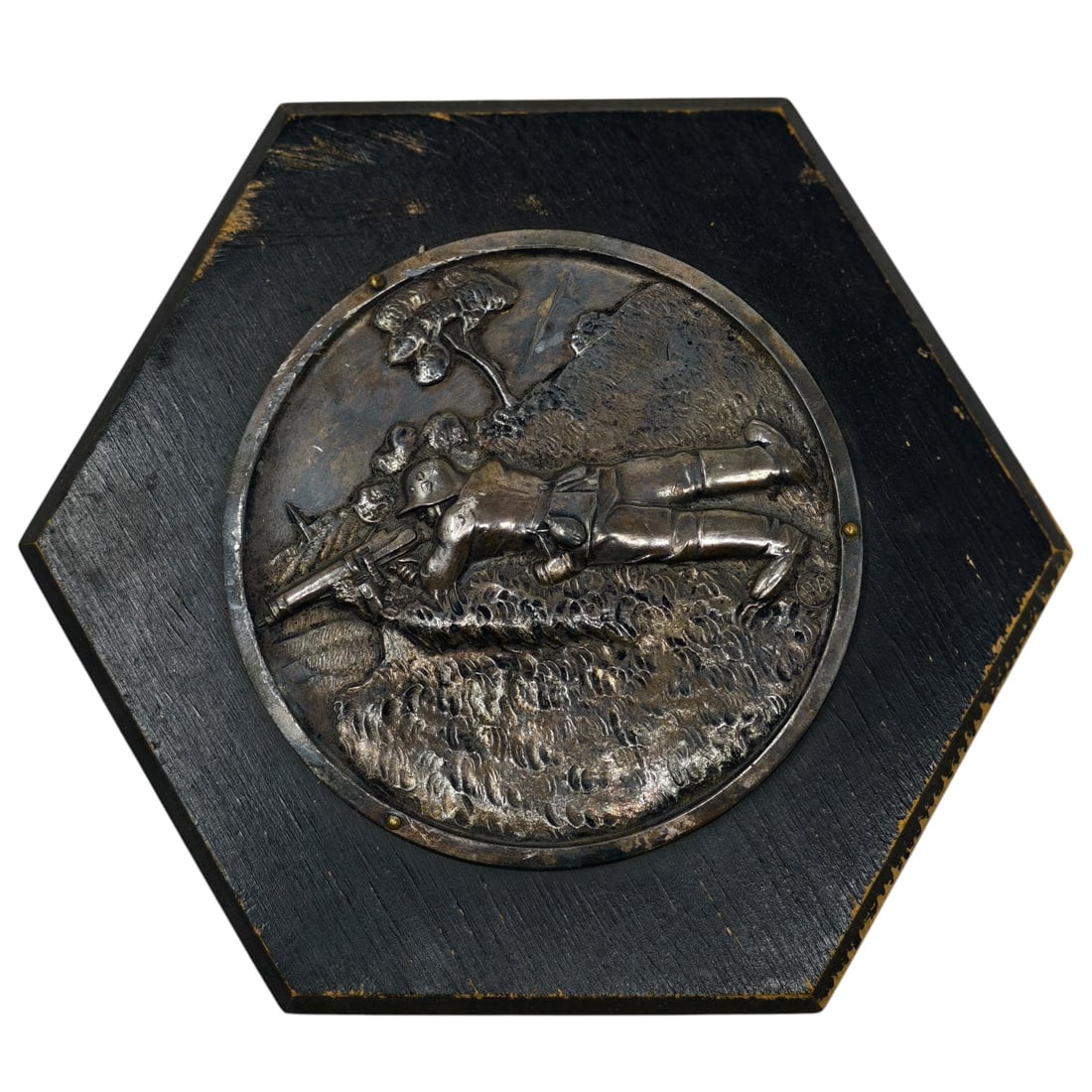 Third Reich Wehrmacht Machine Gun Shooting Plaque: A hexagonal wooden wall plaque with a circular repoussé medallion in what appears to be metal depicting a prone soldier operating a machine gun in a trench or field setting, with foliage and what app