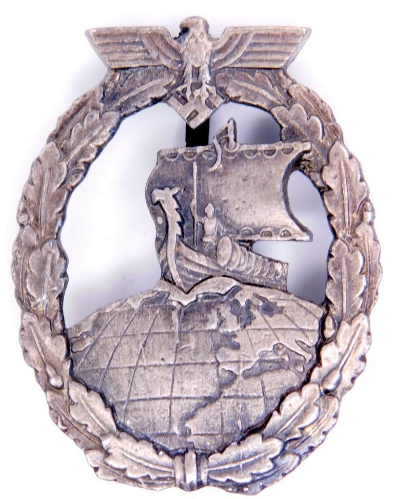 German WWII Naval Kreigsmarine Auxiliary Cruiser Badge: Brass construction. Front shows a Viking ship in the center with a German eagle clutching a swastika in his talons at the top. Both are surrounded by an oval oak leaf wreath. Wide vertical pin back wi
