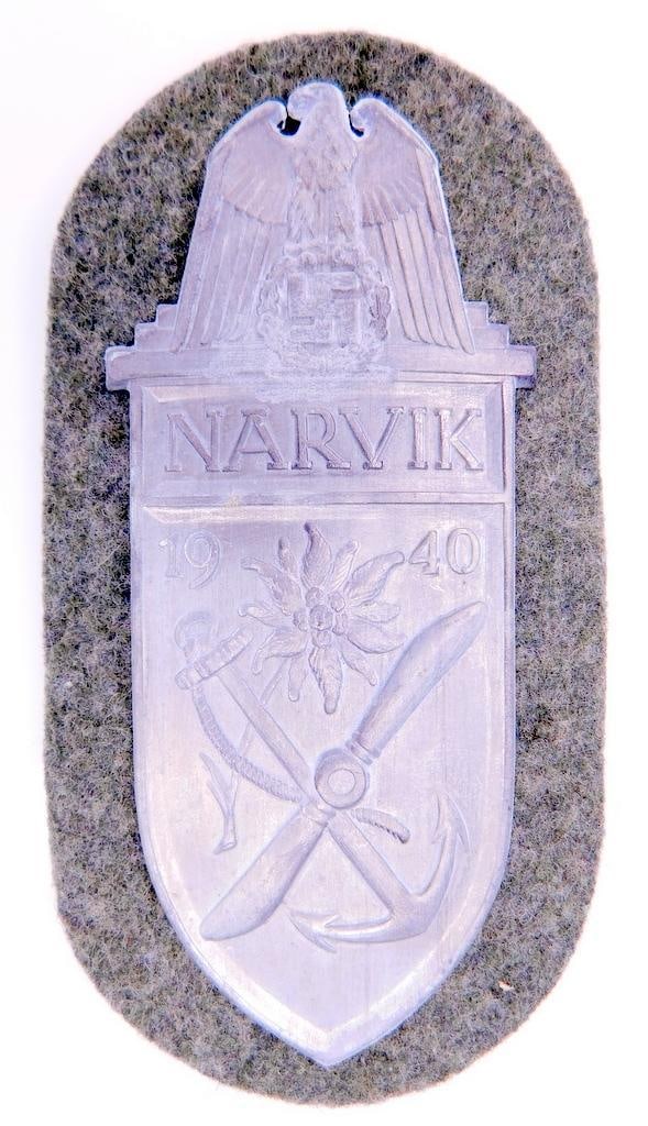 German WWII Army NARVIK 1940 Sleeve Shield: One piece die struck stamped nickel construction. Front shows an edelweiss in the center with a crossed propeller and anchor below. German eagle clutching a swastika in his talons at the top. It reads