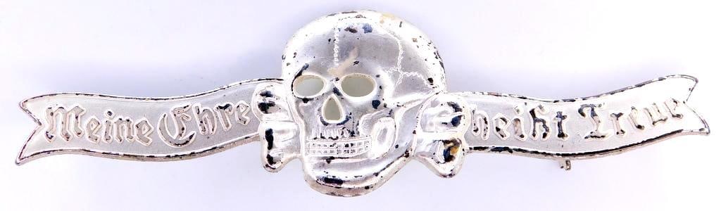 German WWII Waffen SS Skull Breast Badge with motto: One piece die struck silver plated nickel construction. Front shows a Waffen SS skull and cross bones in the center. It reads Meine Ehre heist Treue (My Honor is Loyalty). Thin horizontal pin back wit