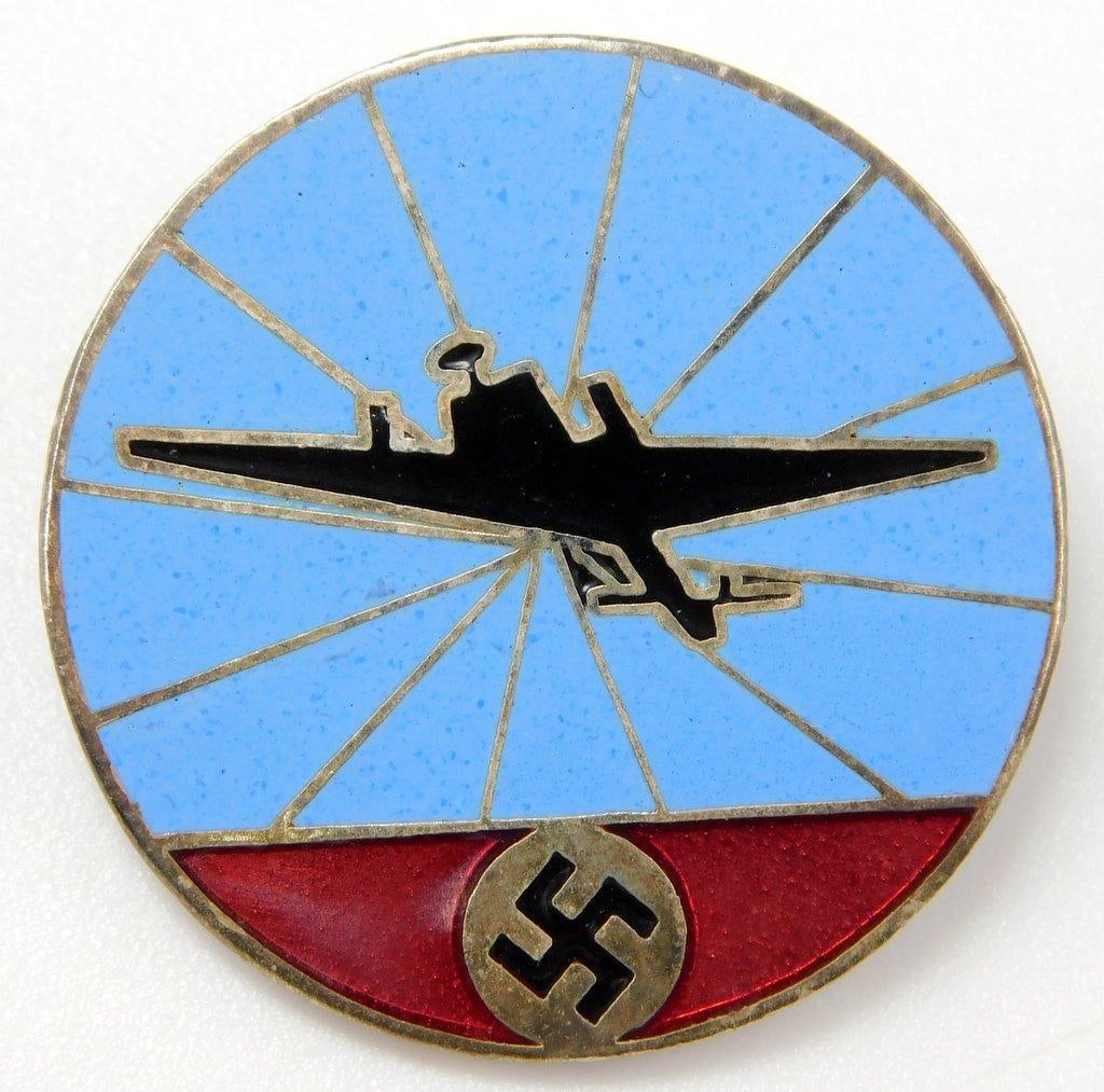 Luftwaffe Air Defense Helper Spotter Badge: One-piece die-struck nickel construction with enamel. Front shows a flying airplane with rays in the background. There is a swastika at the base. Thin horizontal pin back with a locking catch. Measure