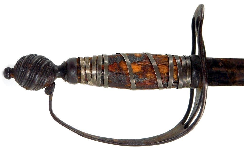 Historic Sword w/ Metal Basket Hilt and Wooden Grip: Antique sword featuring a metal basket hilt and a wooden grip wrapped with metal bands. The blade shows signs of age and wear consistent with age and use. The pommel has a spiral design, and the 