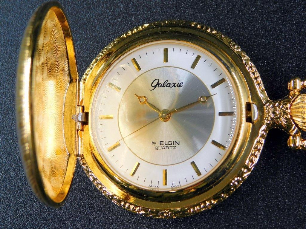 Elgin Galaxie Quartz Pocket Watch with Chain and Original Box: Elgin Galaxie Quartz pocket watch featuring a gold-tone case with a vertical striped design, complemented by a decorative crown and matching gold-tone chain with clip Includes the original