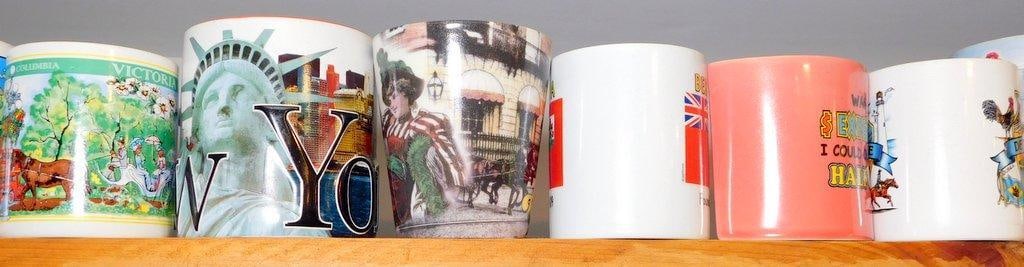 Six Souvenir Coffee Mugs w/ Various Themes: Six souvenir coffee mugs featuring a variety of designs and themes The collection includes imagery of landmarks such as the Statue of Liberty, cultural scenes, international flags, and humorous