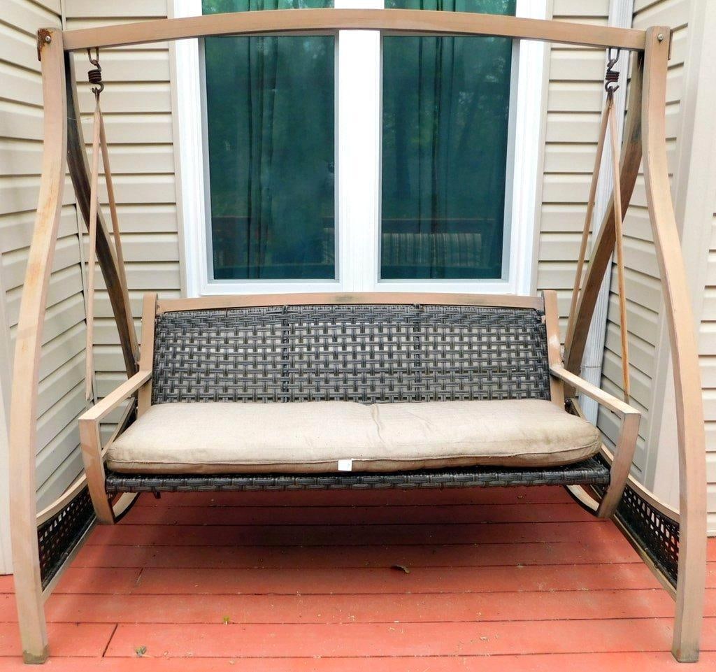 Outdoor Swing w/ Wicker Frame and Sunbrella Cushion: Outdoor swing featuring a beige metal frame with visible rust and wear The seat and backrest are constructed of woven wicker material Cushions included, featuring Sunbrella fabric Signs of weather