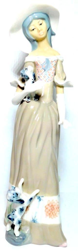 Casades Porcelain Figurine of Woman w/ White Hat and Dogs: A porcelain figurine by Casades, made in Spain, featuring a woman wearing a white hat and gloves, a beige dress with floral patterns, and holding a small dog Another dog is depicted at her feet, which