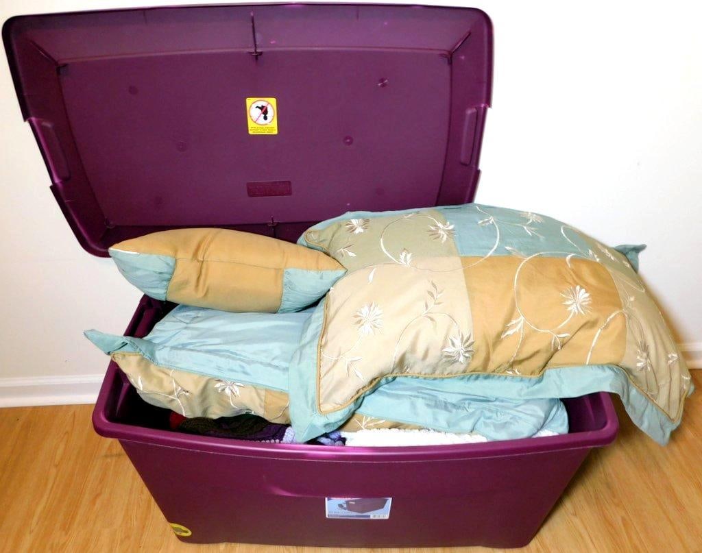 Purple Bin Containing Pillows, Blankets, and Crochet Items: Large purple storage bin containing various home textiles including two decorative pillows with floral embroidery, a set of crocheted blankets in multiple colors, a white knit blanket, and two Happy