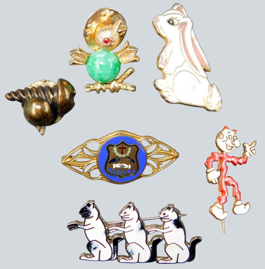Vintage and Contemporary Pins and Brooches Featuring Various Designs: Six pins and brooches, including a whimsical cartoon character pin marked M.R., a white bunny pin made by Pinnacle Designs, a pin with a design of three cats, a shield-shaped blue emblem pin, a bird
