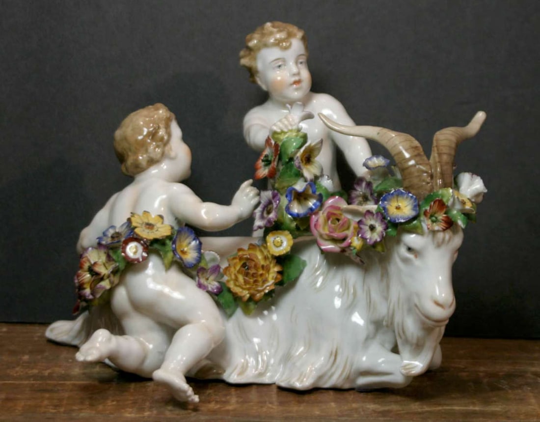 antique von Schierholz porcelain Cherub and goat statue: figural statue grouping in glossy porcelain depicting a pair of full figured brown curly haired, blue eyed nude or naked Cherubs or Putti draping a flower chain around a horned and long haired g