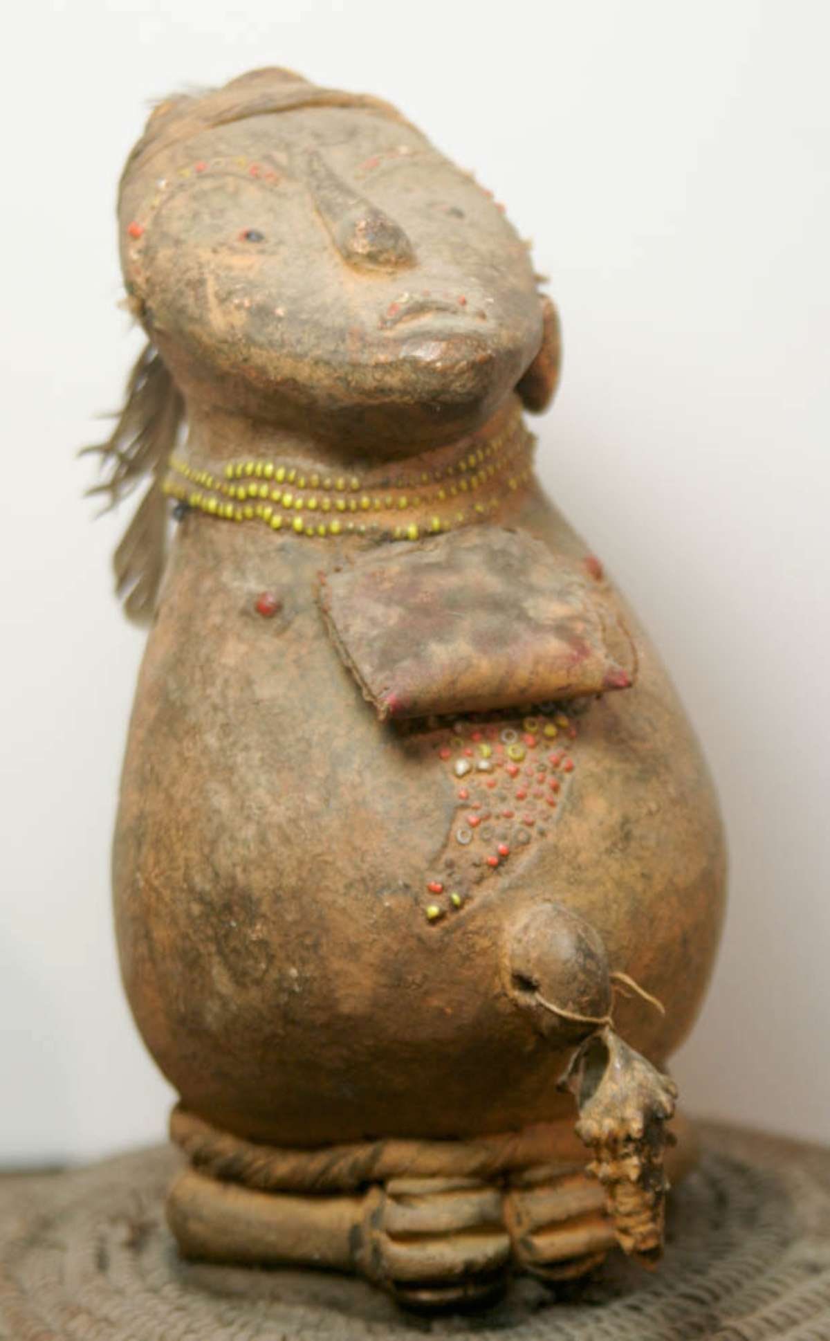 antique African fertility totem: vintage or antique moulded clay or pottery human figure (likely a male) adorned with shells, feather and tiny colored trade seed beads and set on a woven disc base, possibly a fertility or shrine piec