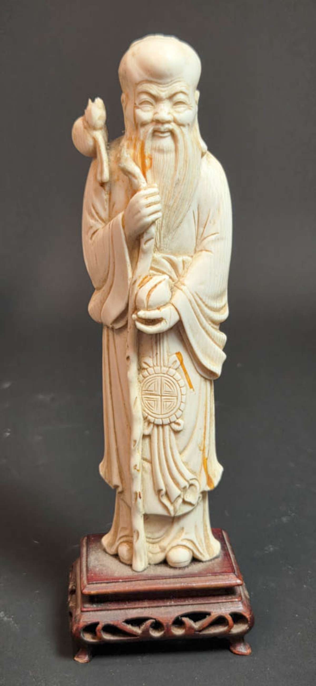 antique Asian carved bone figural statue: on carved wooden stand, this is a beautifully detailed carved antique bone figurine of a man (Wise man, Saint or Deity?) with a high forehead and holding a tall staff and peach or other fr