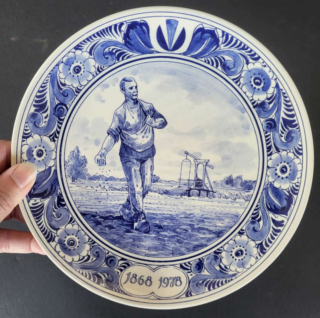 antique porcelain Delft plate with farmer: 1868 to 1978 110 year commemorative anniversary pottery or porcelain plate with white background and blue hand painted floral or flower design border and the center depicting a Dutch (from Holland&com