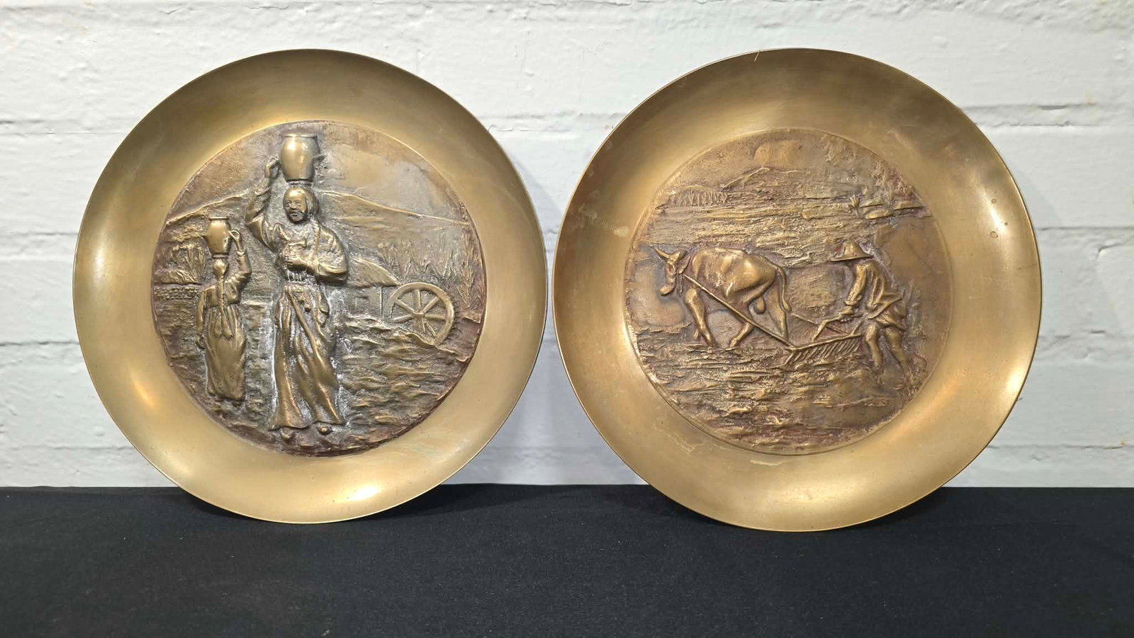 Vintage Brass Wall Plates with Rural Scenes: Decorative pair of brass relief plates featuring detailed pastoral and rural genre scenes in raised relief.One plate depicts a farmer plowing a field with oxen, while the companion plate shows figures