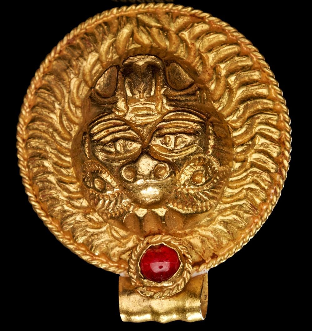A GREEK GOLD & GARNET AMULET DEPICTING A FACE, HELLENISTIC PERIOD, CIRCA LATE 4TH CENTURY B.C.: A GREEK GOLD & GARNET AMULET DEPICTING A FACE, HELLENISTIC PERIOD, CIRCA LATE 4TH CENTURY B.C. Struck in rich, warm-toned gold, this compelling Hellenistic amulet takes the form of a circular me