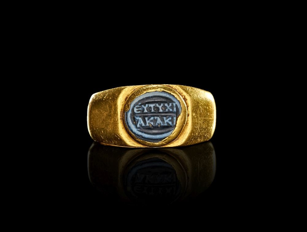 A ROMAN GOLD & NICOLO FINGER RING WITH GREEK INSCRIPTION, CIRCA 3RD CENTURY A.D.: A ROMAN GOLD & NICOLO FINGER RING WITH GREEK INSCRIPTION, CIRCA 3RD CENTURY A.D. Of high-karat gold, this Roman finger ring is formed with a broad, substantial hoop and a raised oval bezel set wit