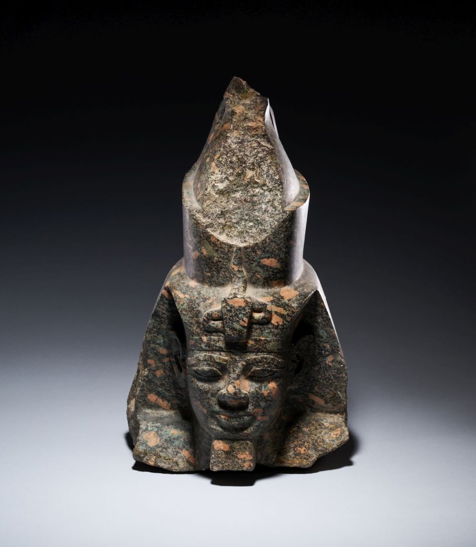 AN EGYPTIAN GRANODIORITE HEAD DEPICTING A KING, POSSIBLY AMENHOTEP III: AN EGYPTIAN GRANODIORITE HEAD DEPICTING A KING, POSSIBLY AMENHOTEP III Carved from mottled granodiorite, this Egyptian royal head preserves the face, crown and broad side elements of the headdress in 