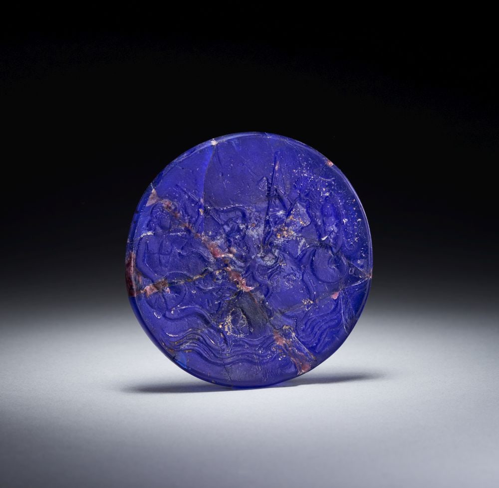 A ROMAN DARK BLUE GLASS DISC DEPICTING A MYTHOLOGICAL SCENE, CIRCA 2ND-3RD CENTURY A.D.: A ROMAN DARK BLUE GLASS DISC DEPICTING A MYTHOLOGICAL SCENE, CIRCA 2ND-3RD CENTURY A.D. Formed in dark blue glass, this circular disc has a compact, medallion-like presence, its rich cobalt colour giv