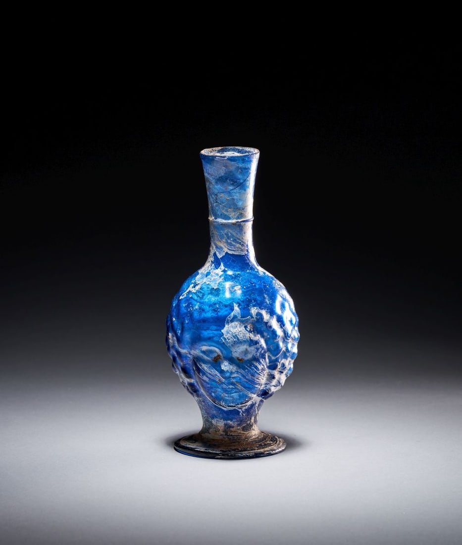 A RARE ROMAN BLUE GLASS JUG WITH A FACE, CIRCA 2ND-3RD CENTURY A.D.: A RARE ROMAN BLUE GLASS JUG WITH A FACE, CIRCA 2ND-3RD CENTURY A.D. Blown in vivid blue glass, this small Roman jug has a tall cylindrical neck, rounded body, applied handle and spreading circular foo