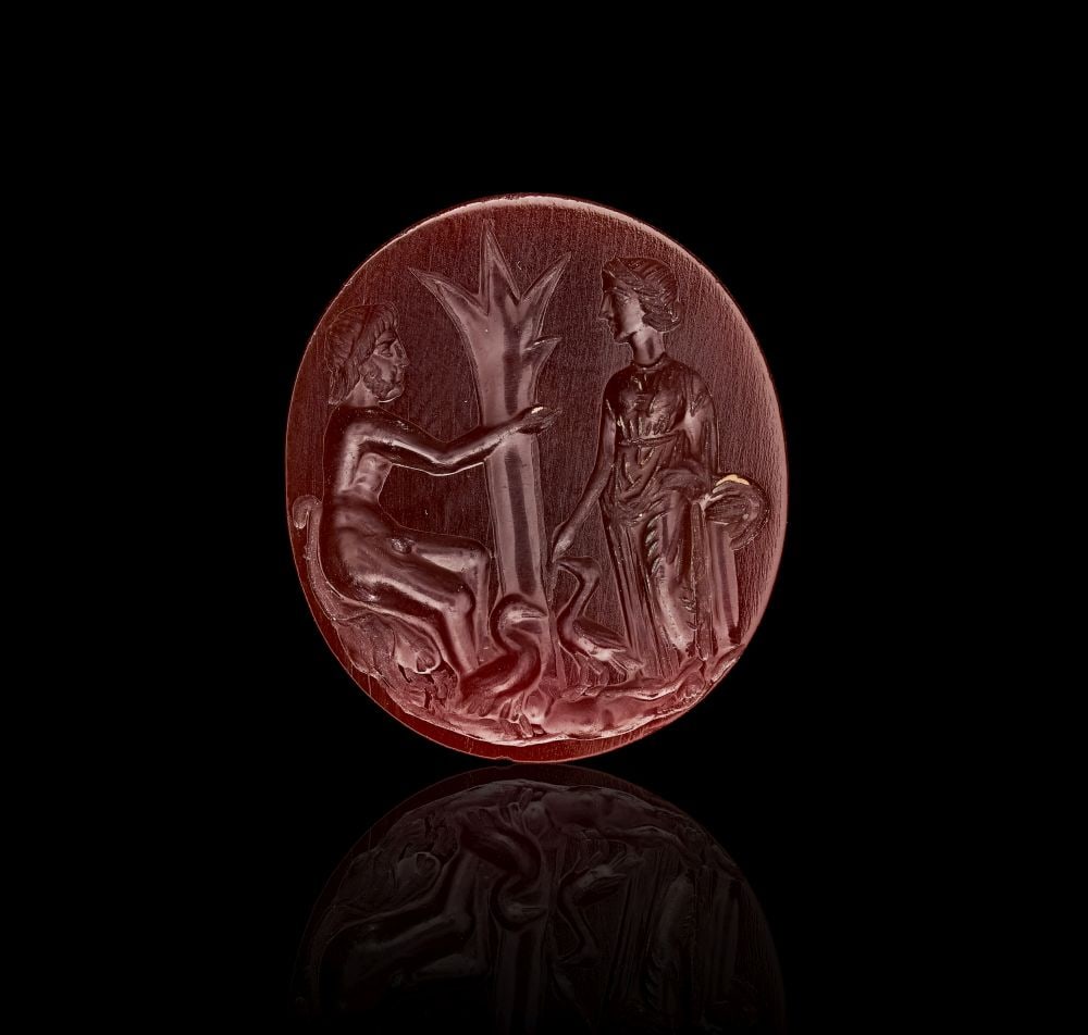 A LARGE ROMAN RED AGATE DEPICTING VENUS & CUPID, CIRCA 1ST-2ND CENTURY A.D.: A LARGE ROMAN RED AGATE DEPICTING VENUS & CUPID, CIRCA 1ST-2ND CENTURY A.D. Carved from a rich red agate, this large Roman intaglio presents a compact mythological scene within an oval field. The 