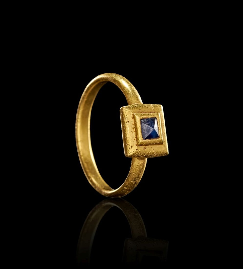 A LATE ROMAN GOLD AND SAPPHIRE FINGER RING CIRCA 3RD-4TH CENTURY A.D.: A LATE ROMAN GOLD AND SAPPHIRE FINGER RING CIRCA 3RD-4TH CENTURY A.D. Formed in gold, this Late Roman finger ring is set with a small square sapphire raised within a stepped rectangular bezel. The pla