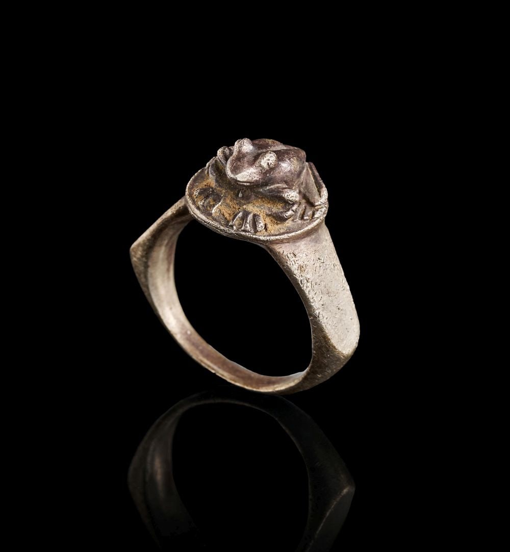 AN EGYPTIAN SILVER FROG RING NEW KINGDOM, DYNASTY XVIII, 1550-1307 B.C.: AN EGYPTIAN SILVER FROG RING NEW KINGDOM, DYNASTY XVIII, 1550-1307 B.C. Formed in silver, this Egyptian ring is centred by a raised bezel modelled as a frog. The hoop is broad and simply shaped, widen