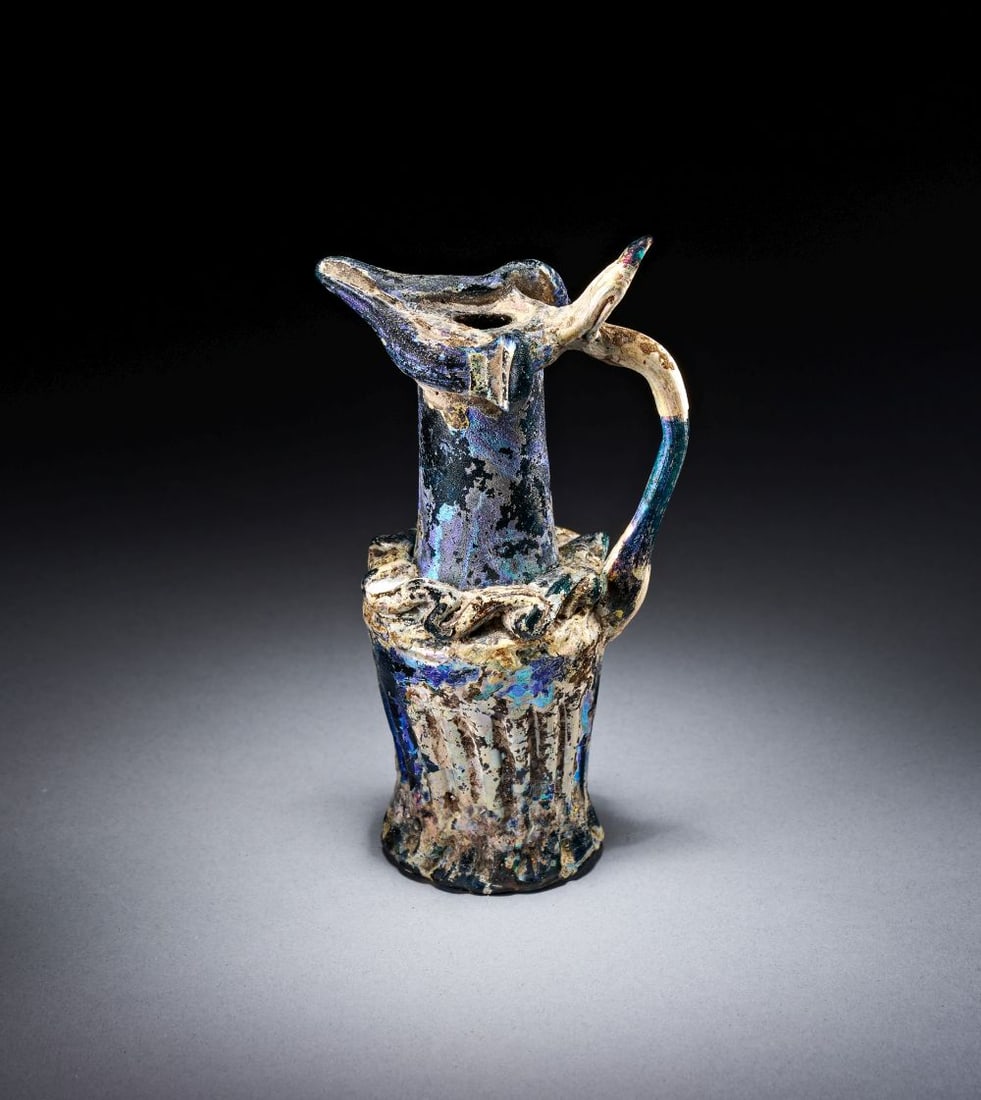 AN UMAYYAD OR EARLY ABBASID GLASS BEAKER JUG MEDITERRANEAN, 8TH/9TH CENTURY: AN UMAYYAD OR EARLY ABBASID GLASS BEAKER JUG MEDITERRANEAN, 8TH/9TH CENTURY Of iridescent glass, the jug is formed with a narrow cylindrical neck rising from a flared lower body, with a high loop hand