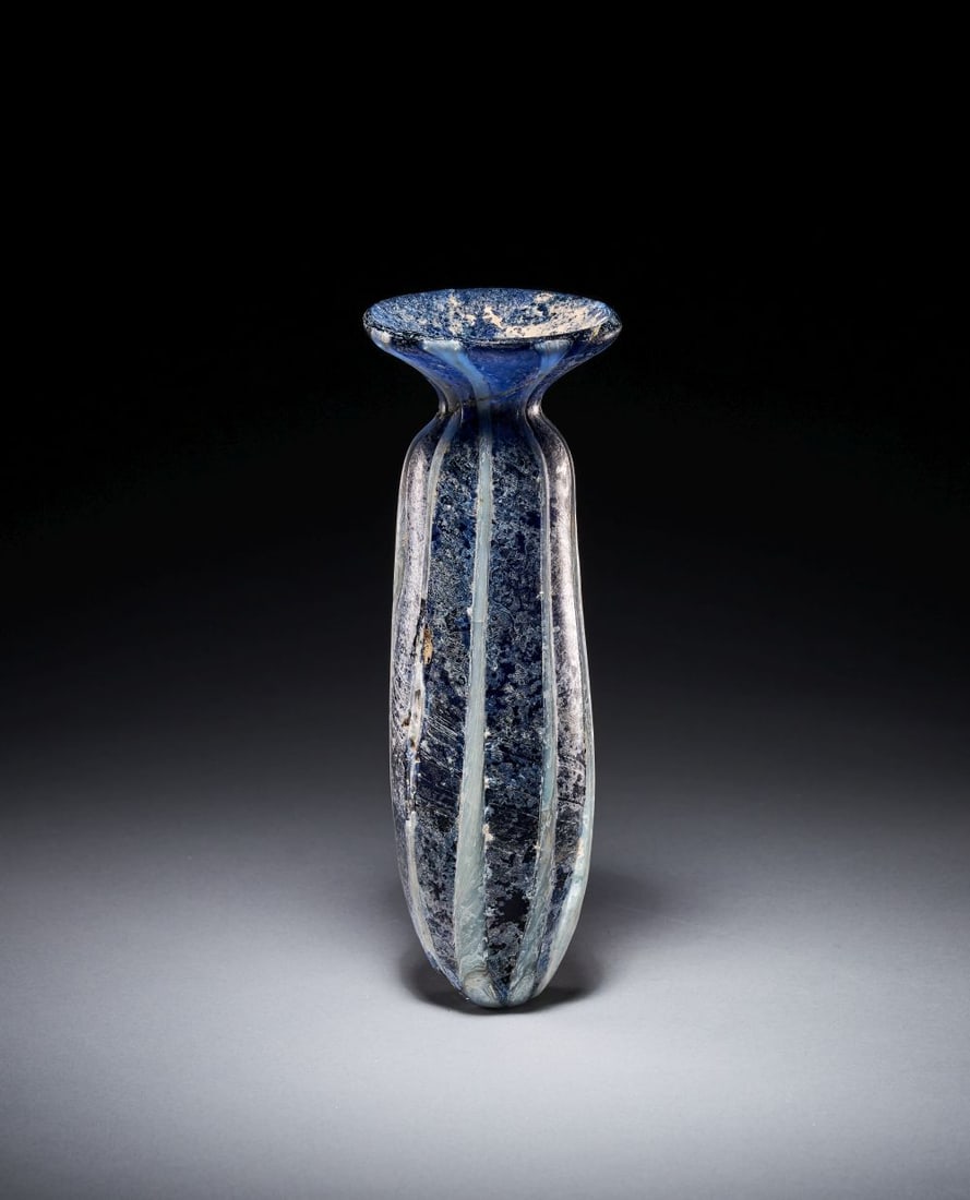 A DARK BLUE MARBLED GLASS ALABASTRON: A DARK BLUE MARBLED GLASS ALABASTRON Of dark blue glass, the vessel is of slender alabastron form, with an elongated tapering body, rounded base and broad flaring mouth. The deep blue ground is enlive