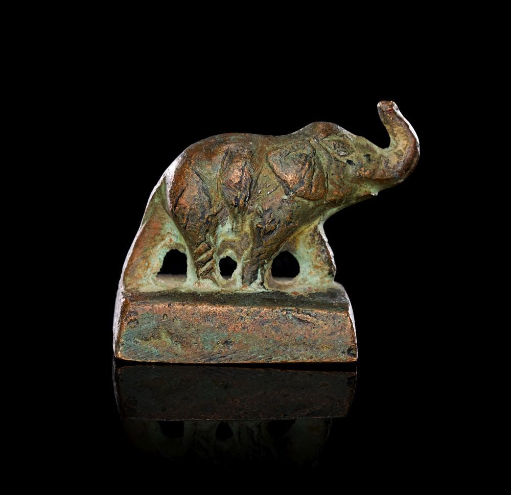 A INDUS VALLEY AMULET SEAL OF A STANDING ELEPHANT WITH HARAPAN INSCRIPTION, CIRCA 2800 B.C. - 1900 B: A INDUS VALLEY AMULET SEAL OF A STANDING ELEPHANT WITH HARAPAN INSCRIPTION, CIRCA 2800 B.C. - 1900 BC A bronze amulet seal modelled as a standing elephant on a rectangular plinth, the animal shown in 