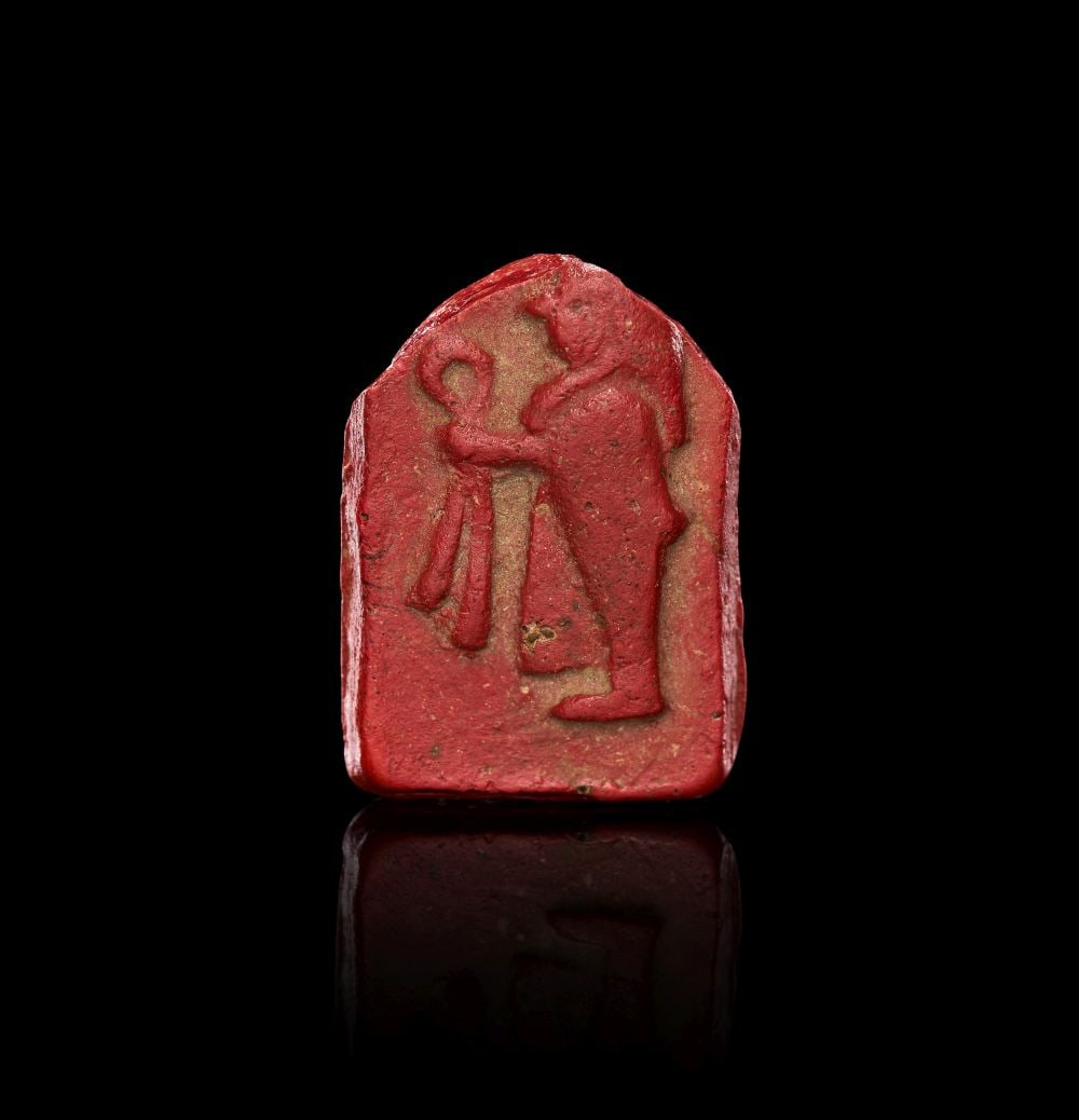 A RED GLASS INLAY OF HORUS HOLDING A TYET, PTOLEMAIC PERIOD, CIRCA 3RD-1ST CENTURY B.C.: A RED GLASS INLAY OF HORUS HOLDING A TYET, PTOLEMAIC PERIOD, CIRCA 3RD-1ST CENTURY B.C. Moulded in opaque red glass, this small inlay depicts Horus standing in profile within a flat-topped shrine-like